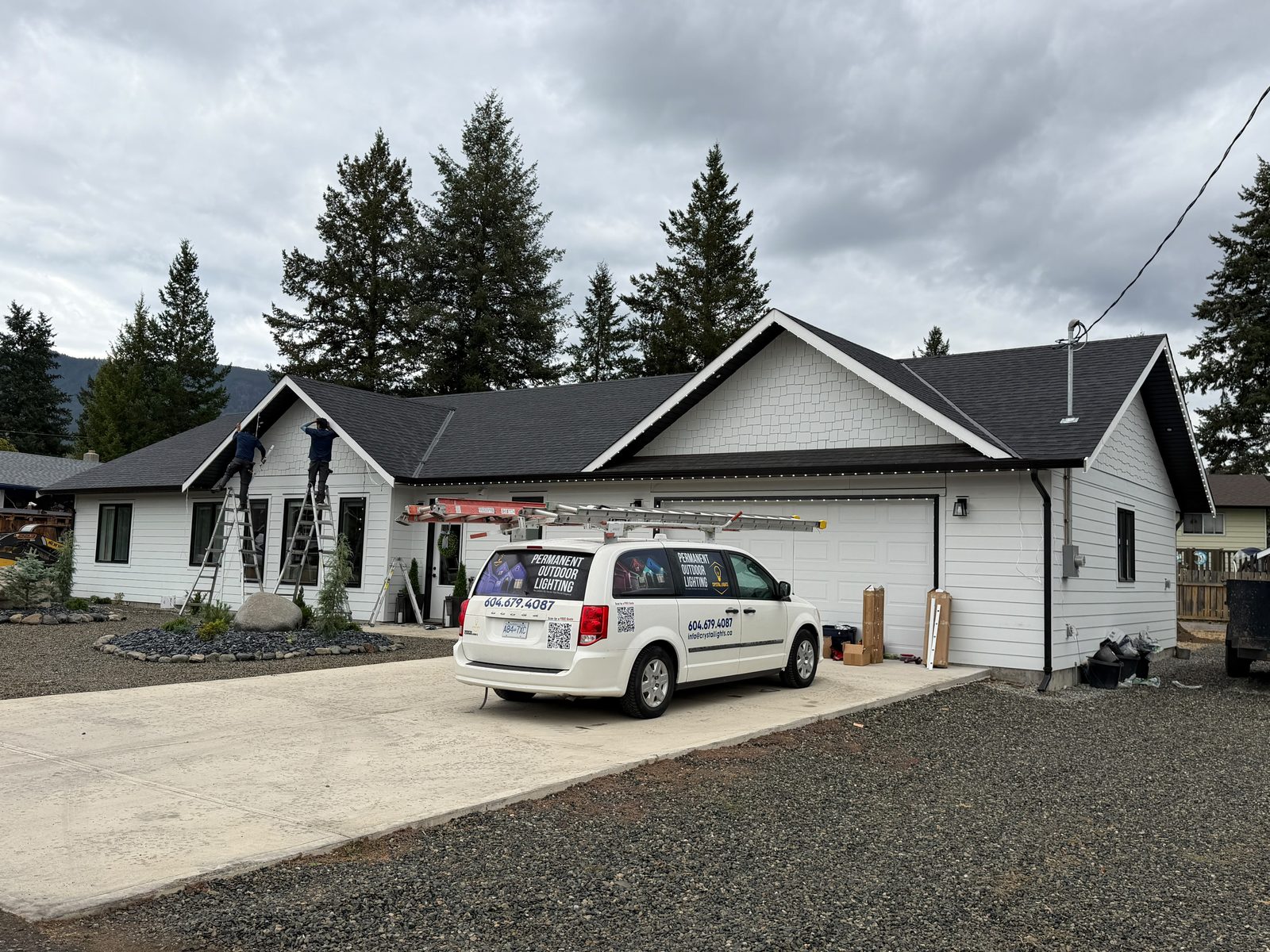 Two Crystal Lights technicians on ladders installing lighting track along a white home's roofline, with the branded company van in the driveway.